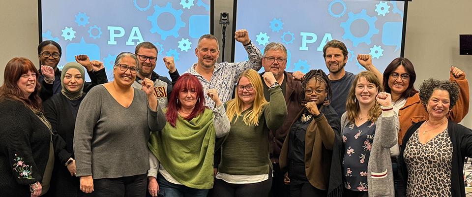 Group photo of the PA bargaining team standing together with solidarity fists raised in front of a backdrop that says PA.
