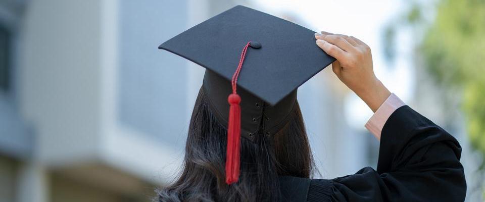 Person in a graduation cap and gown, seen from behind, adjusting their cap with a red tassel, with blurred campus buildings in the background.