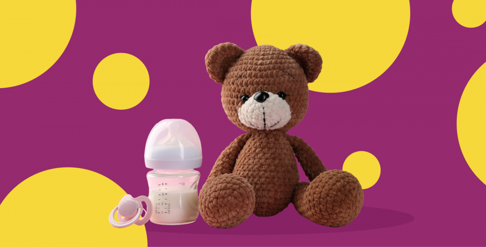 Teddy bear and baby bottle of milk in front of a purple background with yellow polka dots.