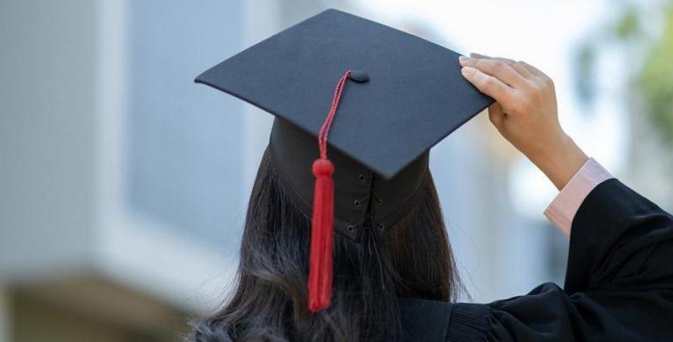 Person in a graduation cap and gown, seen from behind, adjusting their cap with a red tassel, with blurred campus buildings in the background.